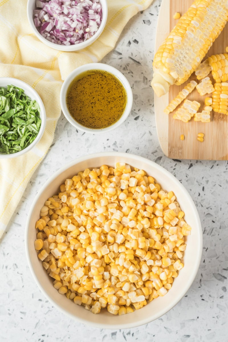 overhead shot of fresh corn salad ingredients: fresh corn, basil, red onion and dressing all displayed in white bowls