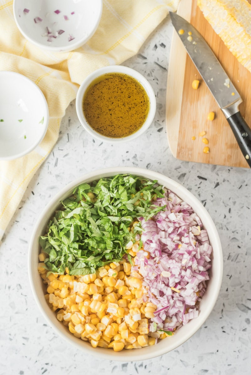 process of making fresh corn salad with corn, onion and basil in a white bowl with dressing displayed on the side