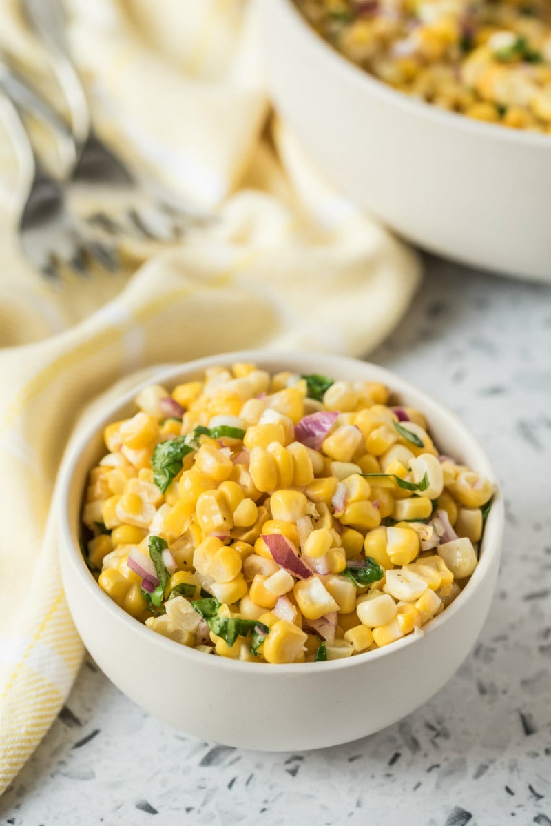 serving of fresh corn salad in a white bowl. larger bowl of corn salad in the background. forks and light yellow napkin displayed too.