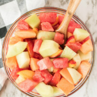 overhead shot of fruit salad in a glass bowl with a wooden spoon. black and white plaid napkin in background