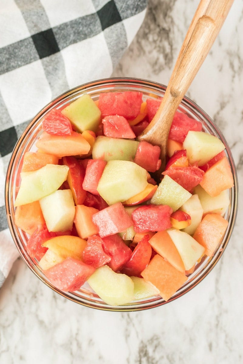 overhead shot of fruit salad in a glass bowl with a wooden spoon. black and white plaid napkin in background