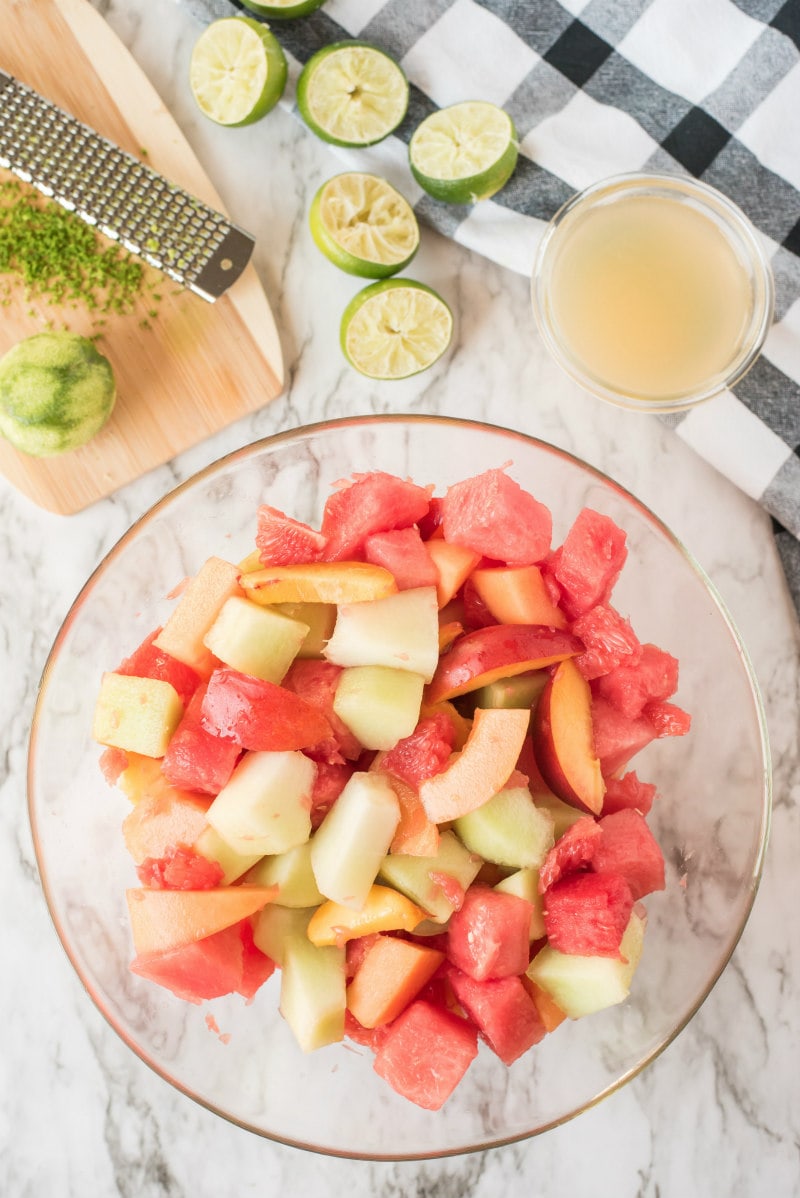 overhead shot of preparation of fruit salad with lime syrup. fruit in glass bowl and zested limes and lime juice in the background