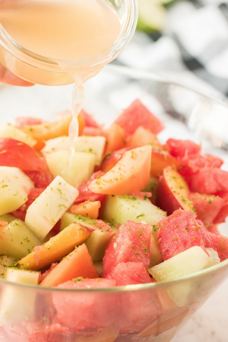 cup pouring lime syrup onto fruit salad in glass bowl