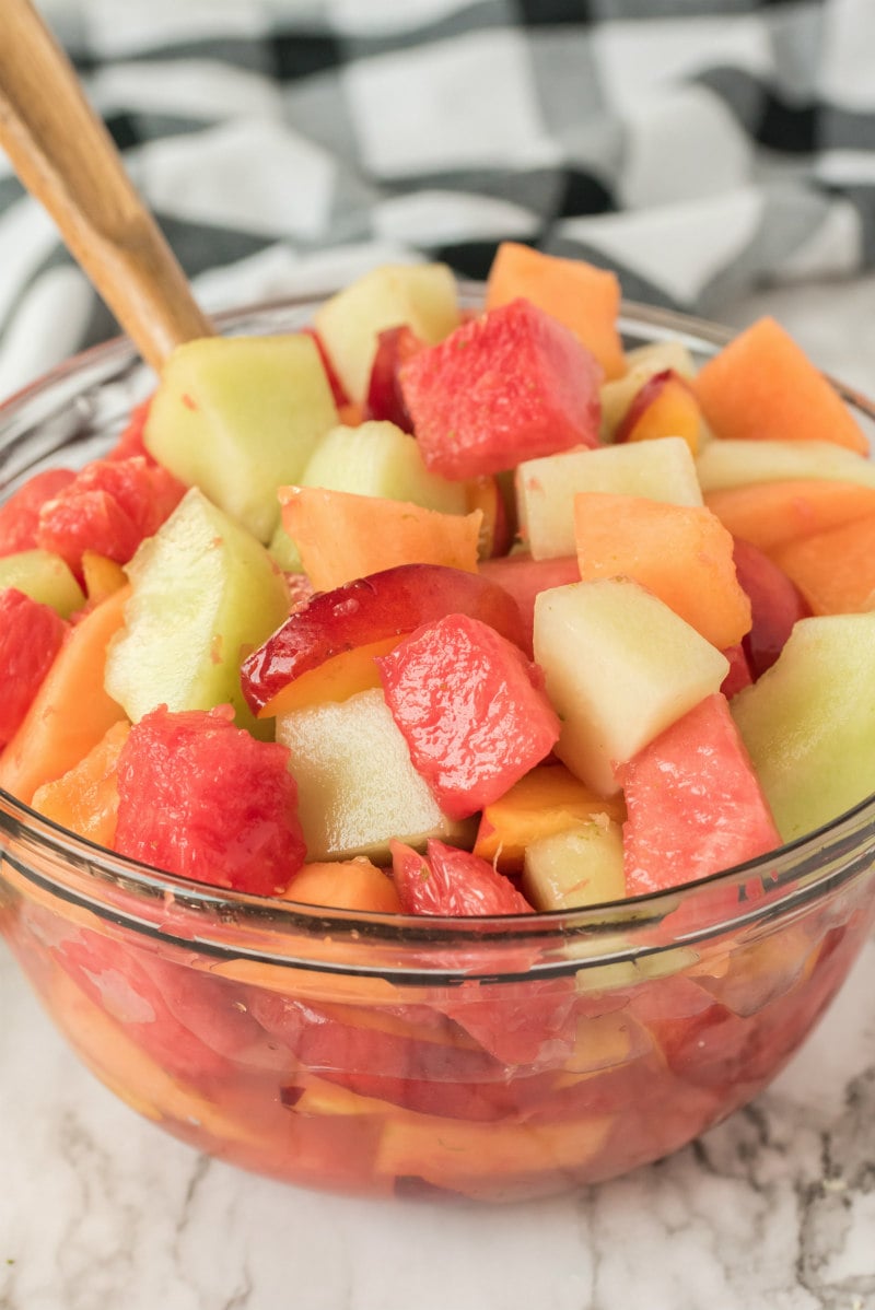 fruit salad with lime syrup in a glass bowl with a wooden spoon. black and white napkin in background.