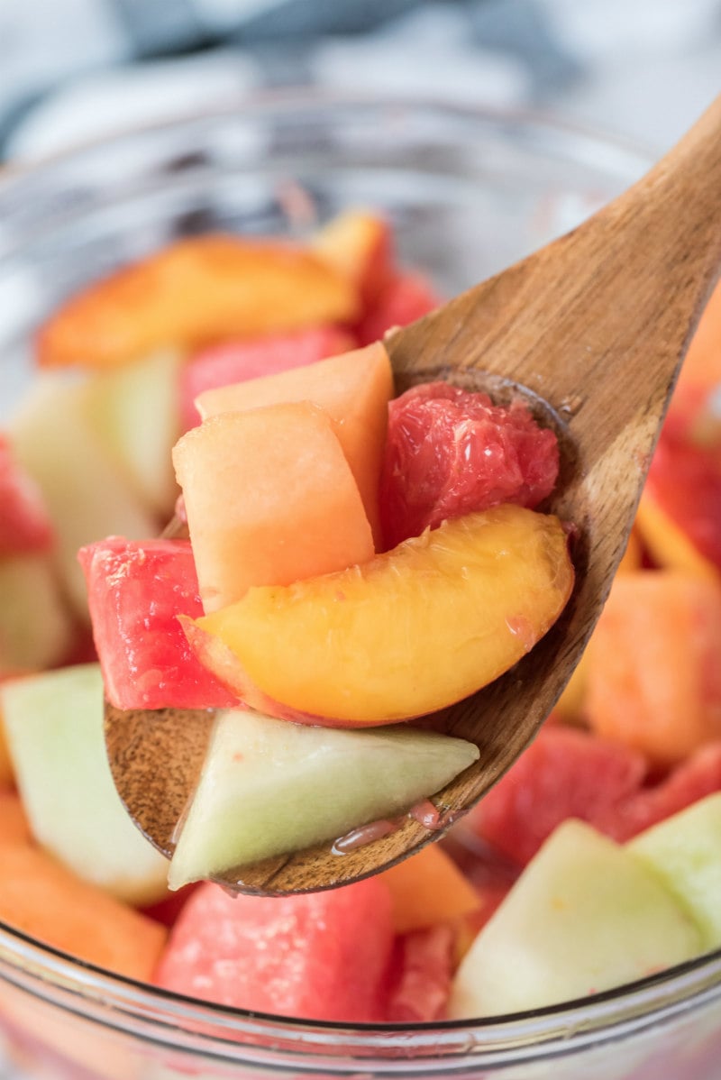 close up of fruit salad in a glass bowl with a wooden spoon