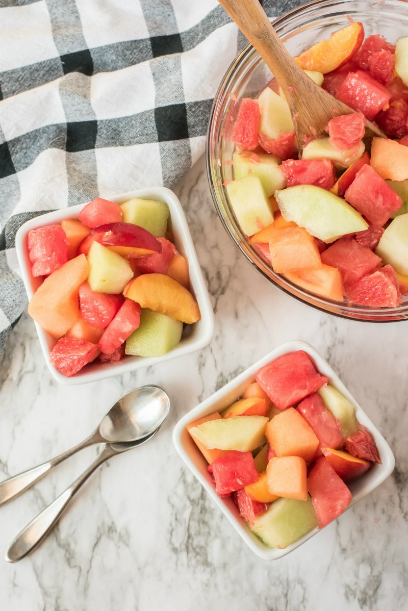 overhead shot of fruit salad with lime syrup in a glass bowl with two servings dished out in white bowls with spoons. black and white checked napkin in background