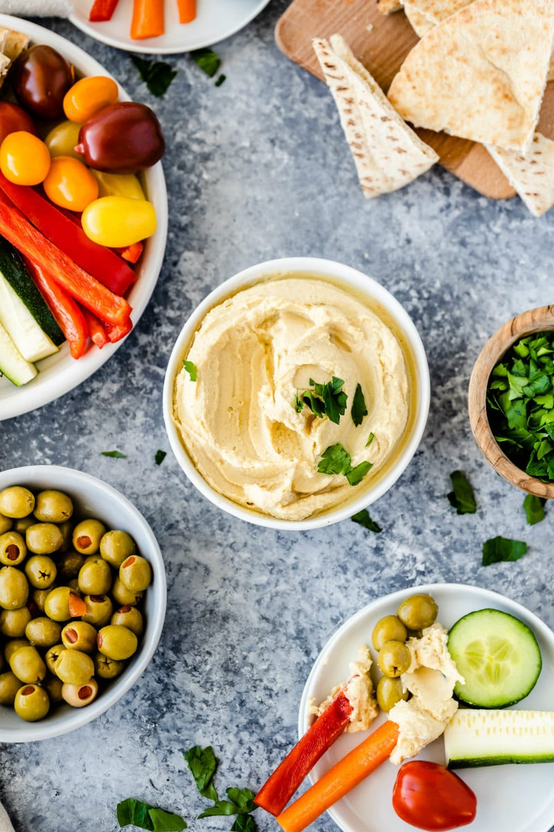 overhead shot of white bowls filled with hummus, olives, tomatoes, peppers, cucumbers and pita bread