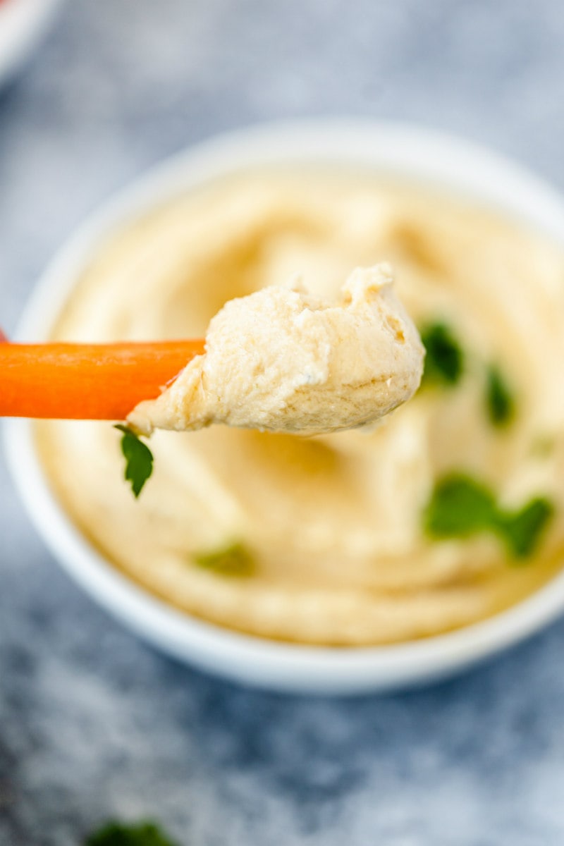 hand dipping carrot into bowl of hummus garnished with parsley