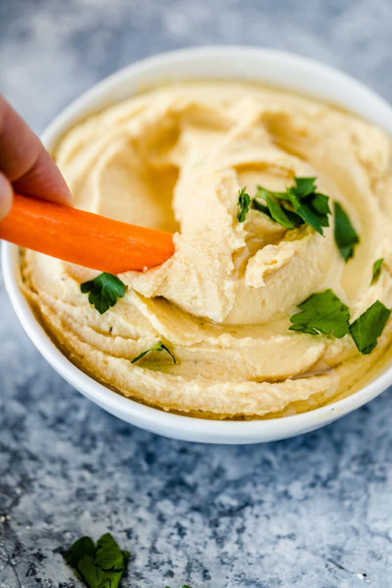 hand dipping carrot into a white bowl of hummus garnished with fresh parsley