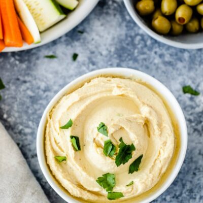 overhead shot of white bowls filled with hummus, fresh vegetables and olives