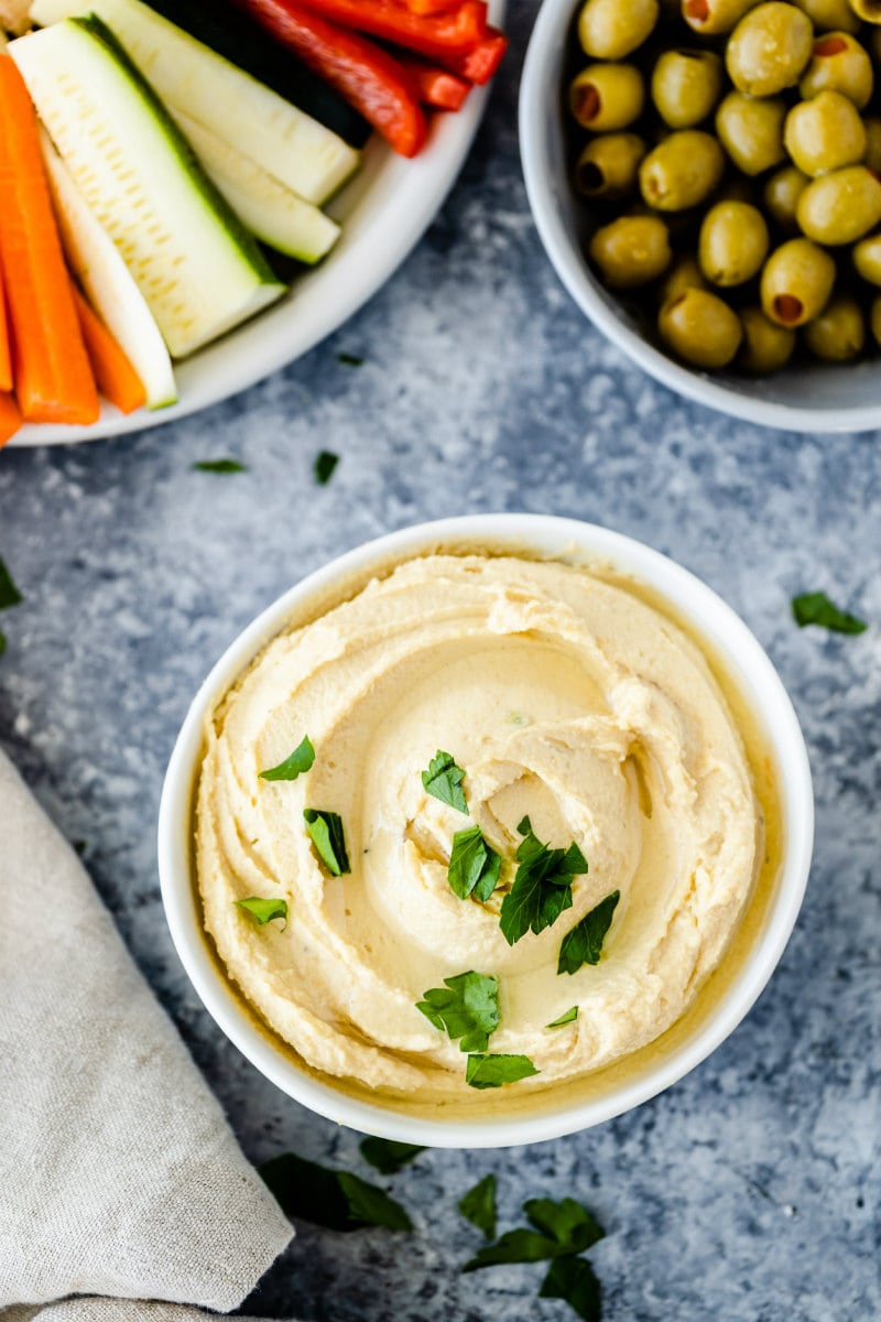 overhead shot of white bowls filled with hummus, fresh vegetables and olives