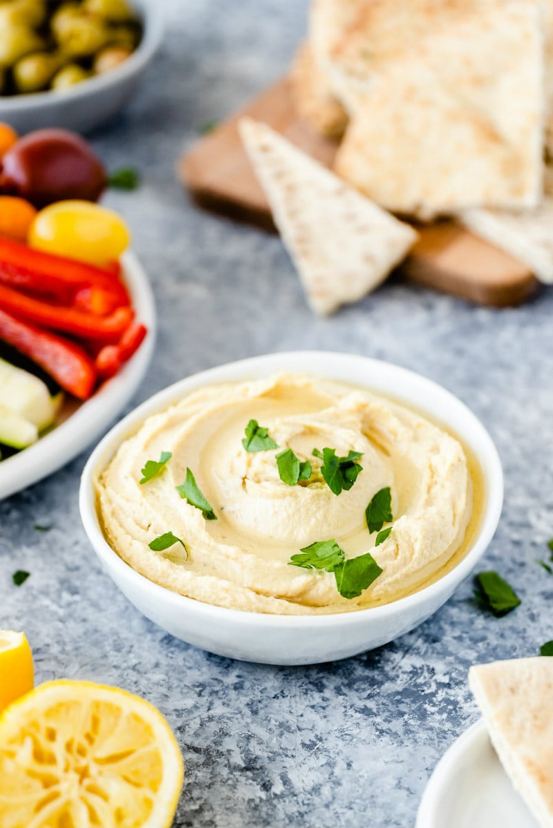 white bowl of hummus surrounded by pita bread and bowls of olives an fresh veggies. squeezed lemon in view too.