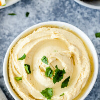 overhead shot of a white bowl of hummus garnished with fresh parsley. peek at fresh veggies and olives in the background