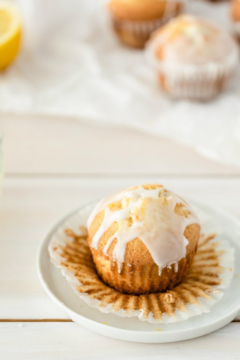 lemon pound cake muffin with glaze unwrapped on a white plate with more muffins and lemon in the background