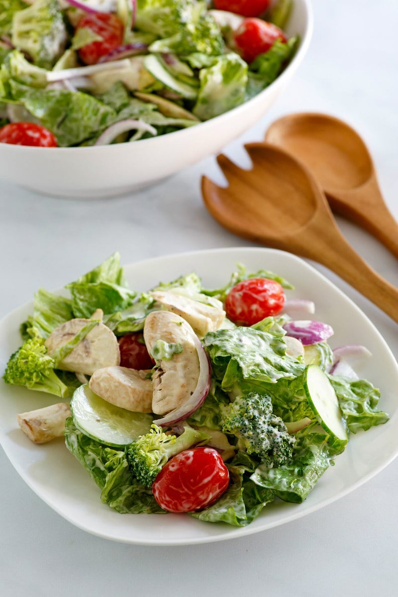 White bowl of Romaine and Broccoli Salad with a white plate of salad on the side. wooden utensils for serving displayed