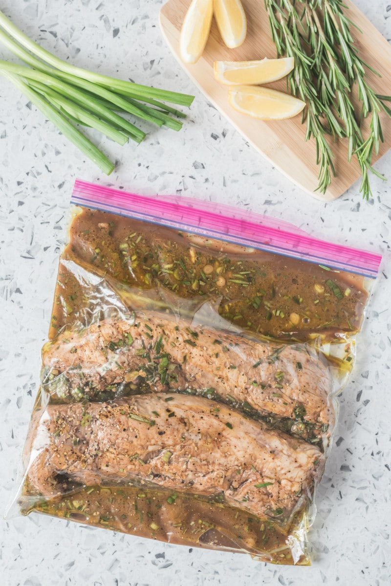 zip plastic bag with two pork tenderloins marinating inside. green onions and lemon in the background