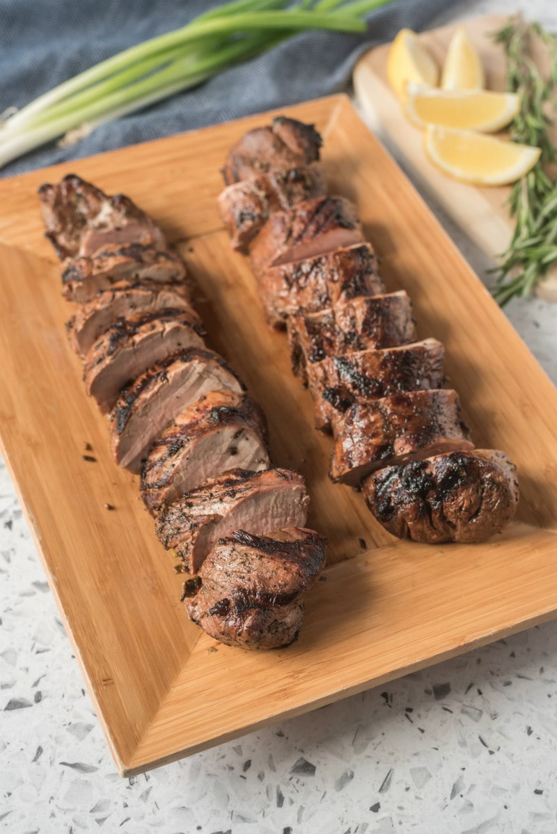 balsamic rosemary grilled pork tenderloin two of them sliced on a cutting board. green onions and lemon wedges in the background