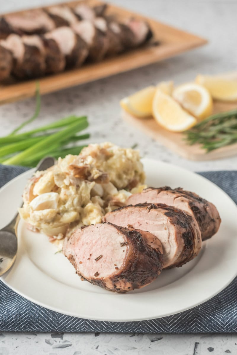 sliced grilled pork tenderloin on a white plate with potato salad with green onions and fresh lemon wedges in the background
