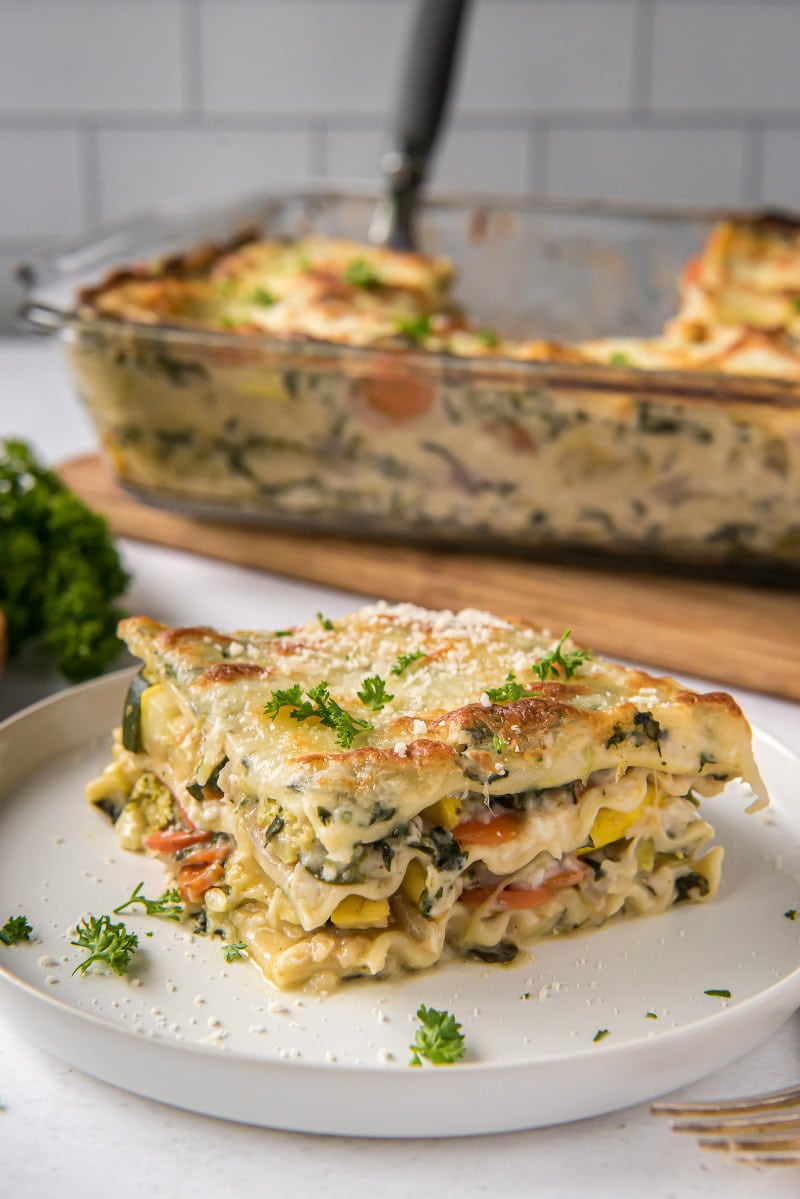 slice of garden vegetable lasagna on a white plate with pan of lasagna in the background