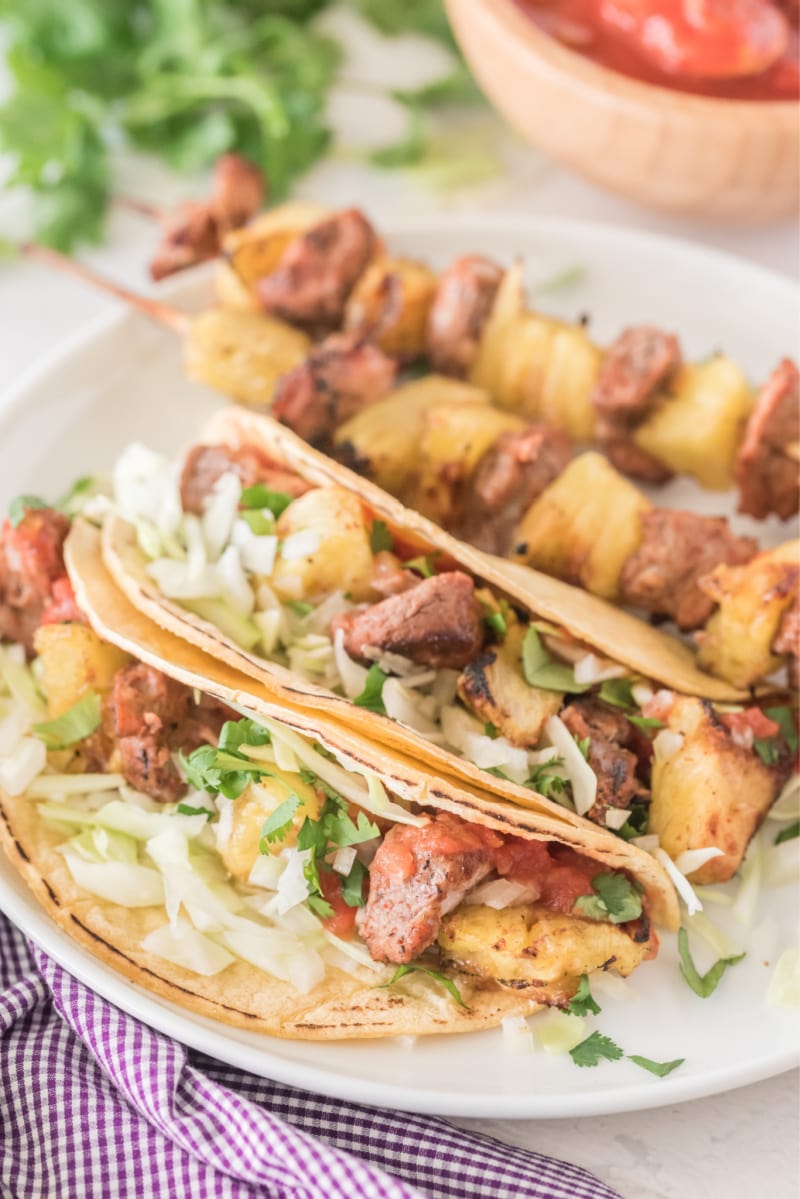 grilled pork and pineapple soft tacos on a white plate with kebabs on plate too