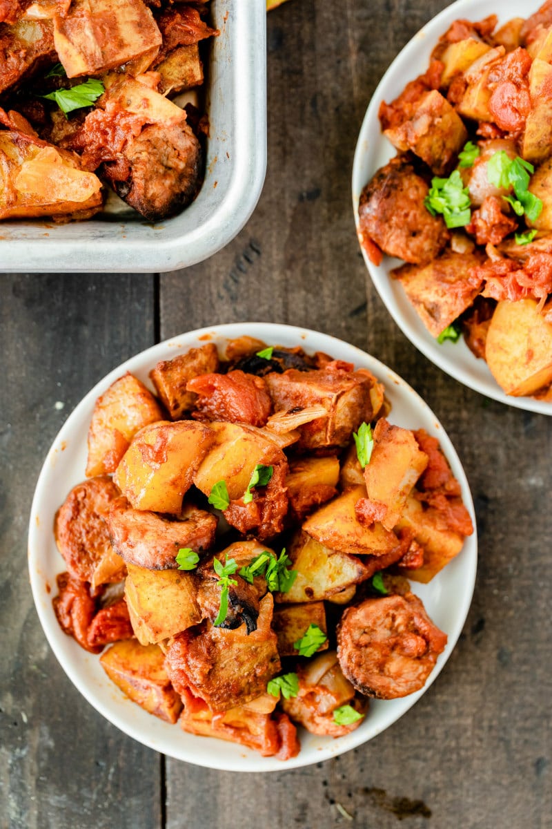 overhead shot of two white bowls of portuguese roasted potatoes garnished with fresh parsley with pan of potatoes in the background