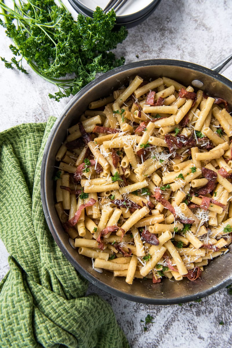 skillet filled with ziti carbonara sitting on a green cloth napkin with fresh parsley on the side