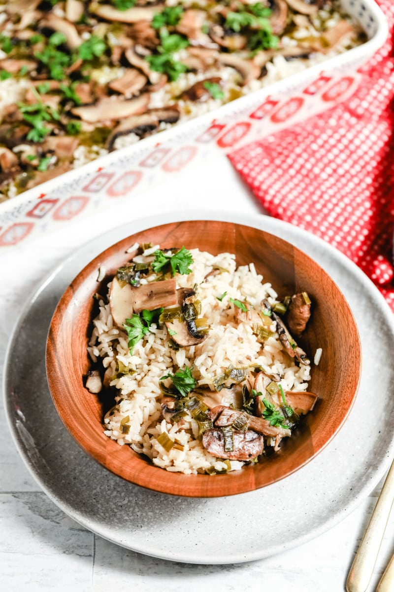 overhead shot of a serving of baked mushroom rice in a wooden bowl wit a red napkin and utensils with casserole dish in the background