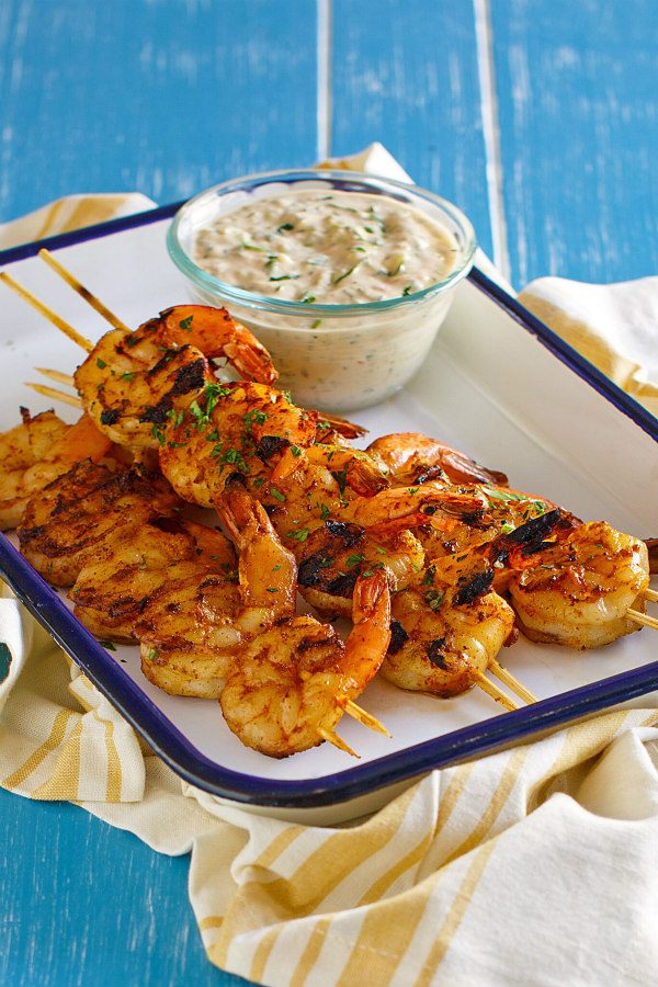 Barbecued shrimp on skewers with a bowl of remoulade sauce served on the side. Set on a white platter with a blue rim. Yellow striped napkin underneath. All set on a blue board background.