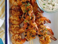 Barbecued shrimp on skewers with a bowl of remoulade sauce served on the side. Set on a white platter with a blue rim. Yellow striped napkin underneath