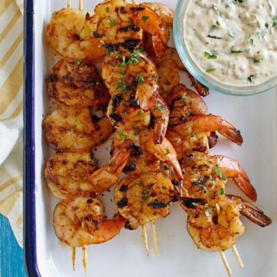 Barbecued shrimp on skewers with a bowl of remoulade sauce served on the side. Set on a white platter with a blue rim. Yellow striped napkin underneath