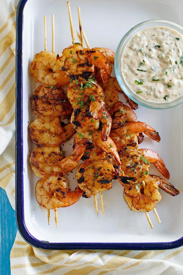 Barbecued shrimp on skewers with a bowl of remoulade sauce served on the side. Set on a white platter with a blue rim. Yellow striped napkin underneath. 