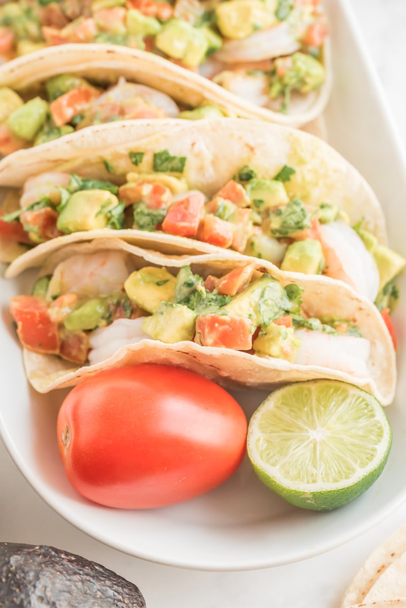 shrimp and avocado tacos on a white platter with tomato and lime