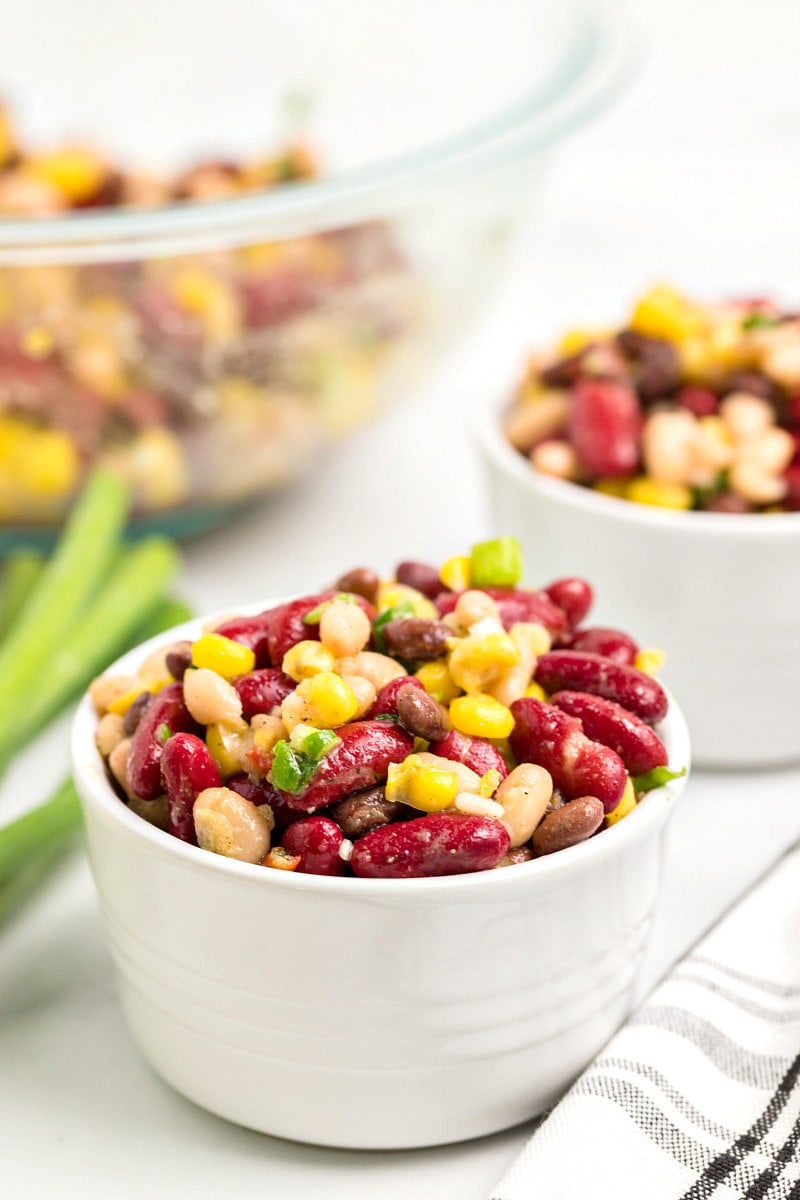 bean salad in a small bowl with the bigger bean salad bowl in the background and green onions garnish