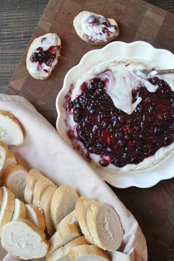 overhead shot of melted baked brie in a white dish with basket of baguette slices on the side and baguette slices with some of the brie and blueberry sauce on them- all sitting on a wooden cutting board