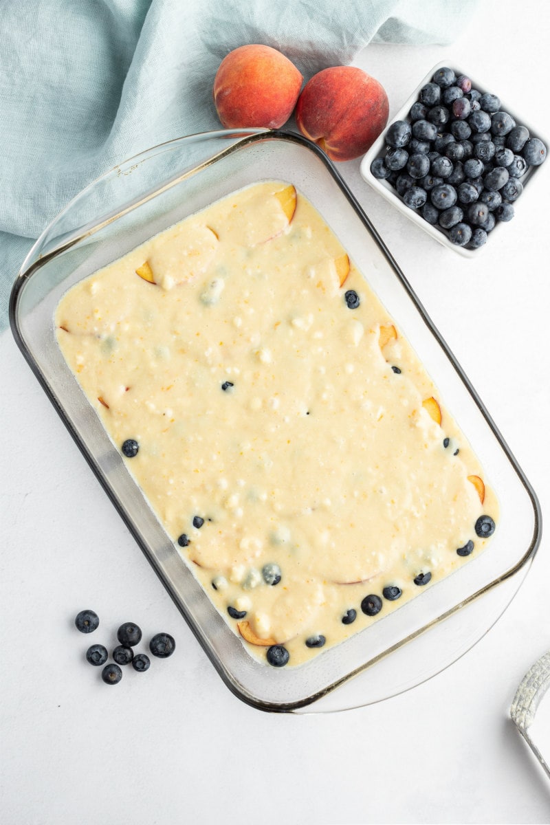 overhead shot of peach and blueberry coffee cake ready to bake in the oven with fresh peaches and blueberries scattered in the background