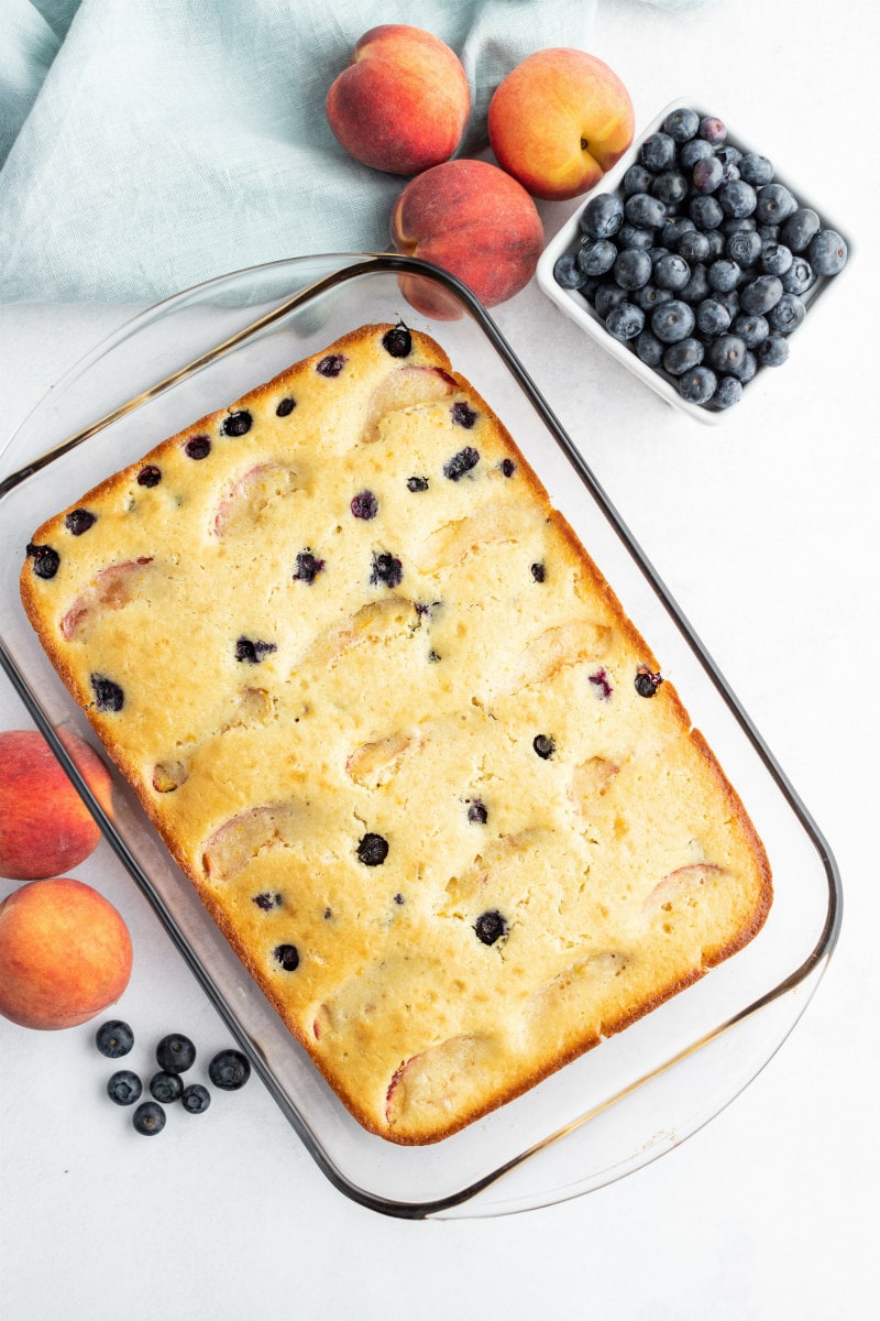 overhead shot of peach and blueberry coffee cake in a pyrex pan with fresh peaches and blueberries scattered in the background