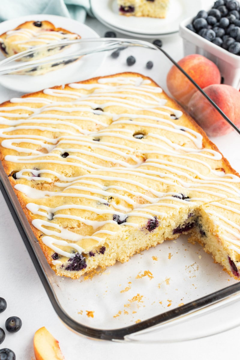 peach and blueberry coffee cake in a pyrex pan with a couple slices cut out of it. peaches and blueberries in the background