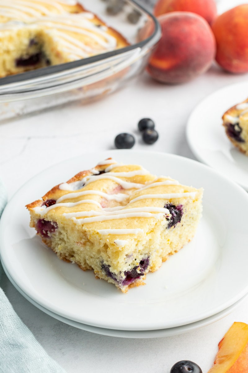 slice of peach and blueberry coffee cake on a white plate with the rest of the coffee cake in the background as well as some fresh peaches and blueberries