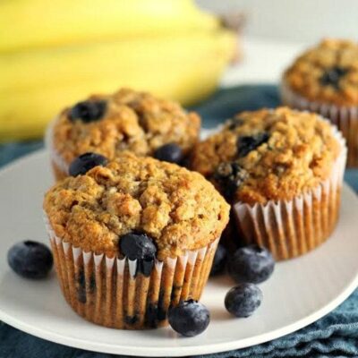 whole wheat banana blueberry muffins (3) on a white plate with fresh blueberries scattered about and bananas in the background