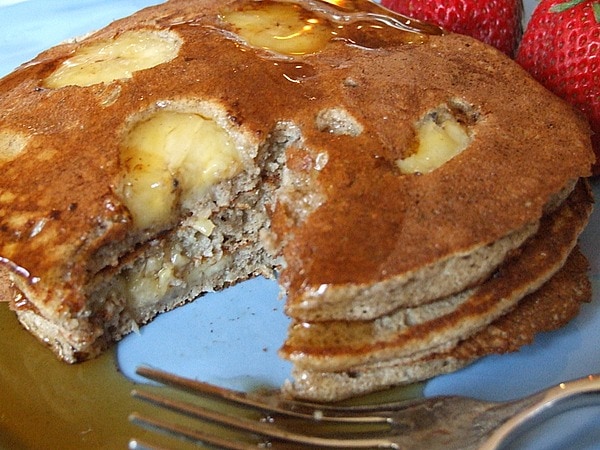 short stack of buckwheat pancakes with bananas and a bite taken out of them on a blue plate with a fork and syrup