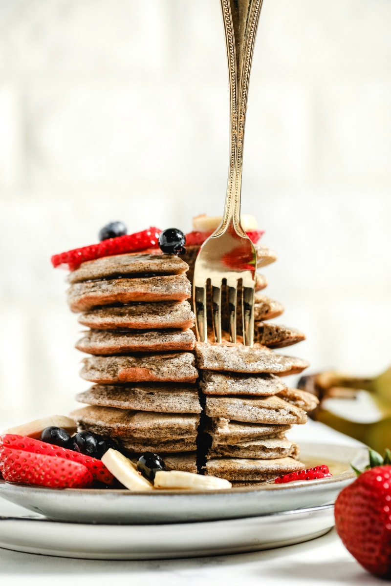 forkful of buckwheat pancakes arranged in a tall stack on a white plate, garnished with fresh berries and bananas