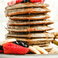stack of buckwheat pancakes on a white plate with syrup drizzling down the sides, garnished with fresh berries and bananas