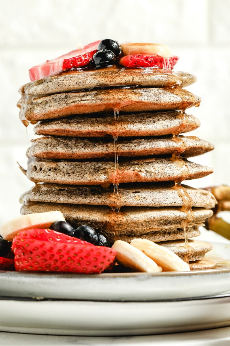 stack of buckwheat pancakes on a white plate with syrup drizzling down the sides, garnished with fresh berries and bananas