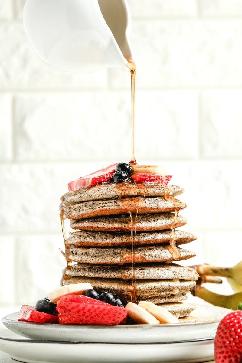pouring syrup onto a stack of buckwheat pancakes on a white plate garnished with fresh berries