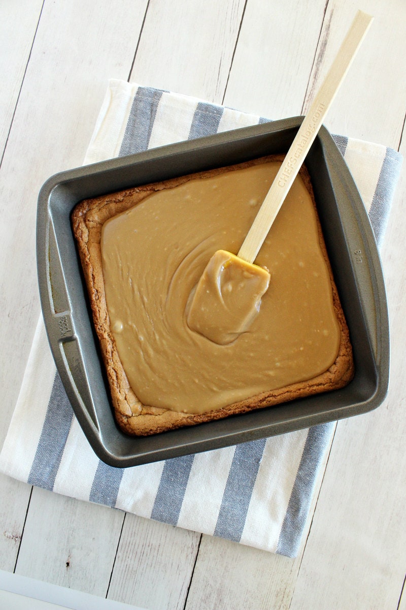 overhead shot of square pan of butterscotch brownies with caramel icing on top with a rubber spatula spreading the icing- on a blue and white striped kitchen towel