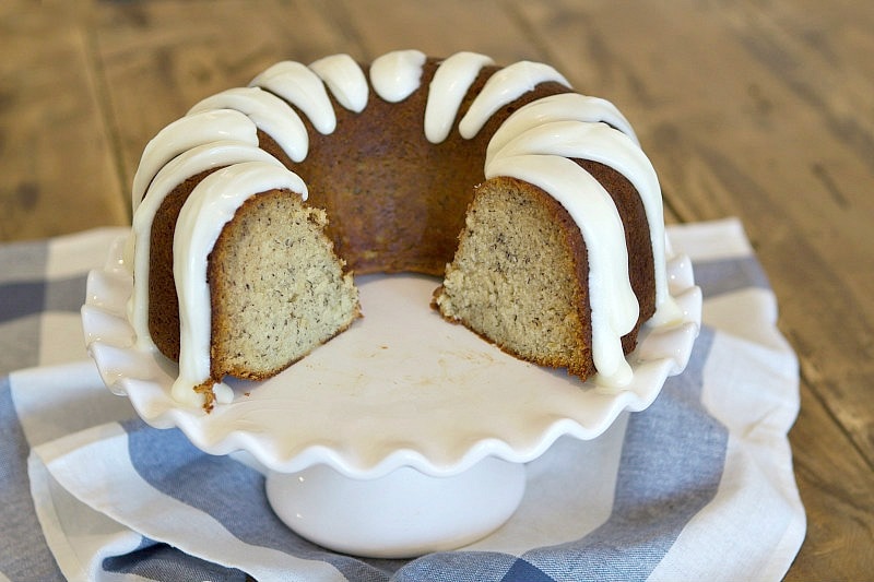 banana bundt cake with cream cheese frosting with a chunk taken out of it displayed on a white cake platter on top of a blue and white striped napkin