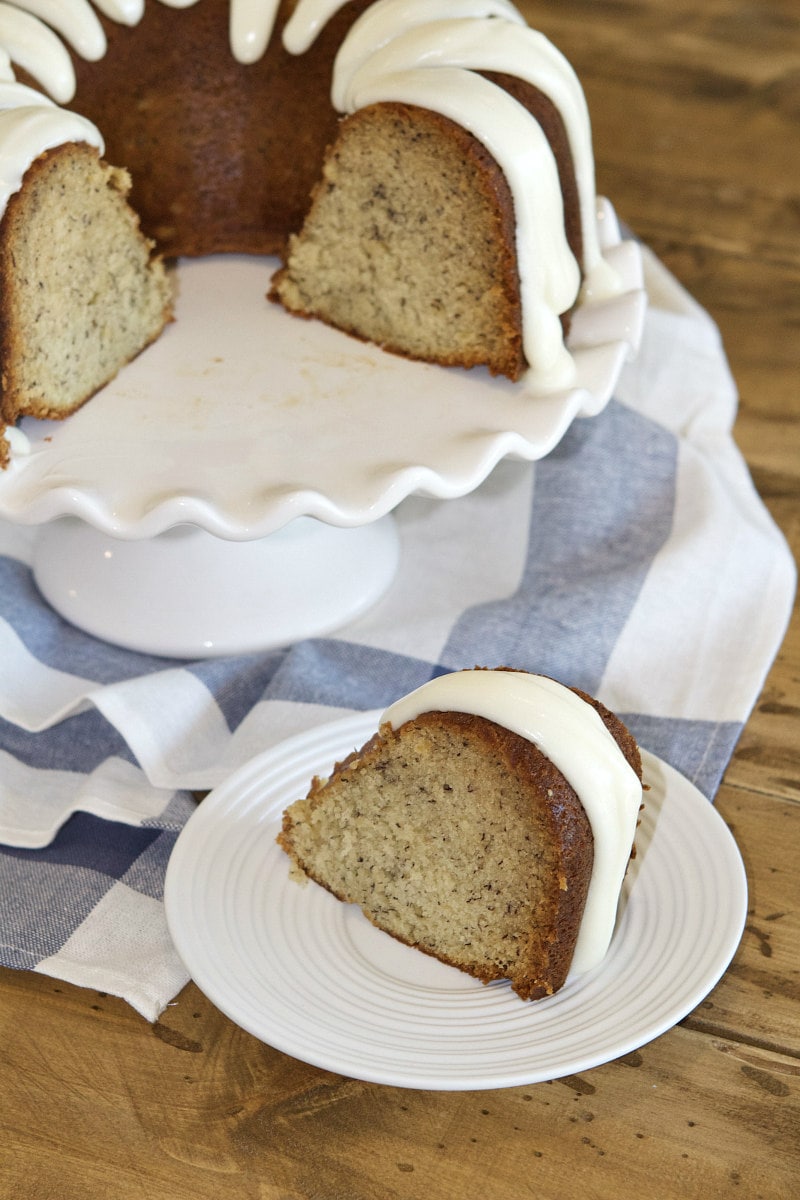 banana bundt cake on a white serving platter with a slice of banana bundt cake on a white plate in front of it. Set on a blue and white striped napkin on a wooden table