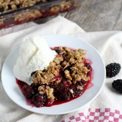 serving of blackberry crisp on a white plate with vanilla ice cream. Set on top of a white and red/white checked cloth napkin. Pan of crisp in the background + fresh blackberries