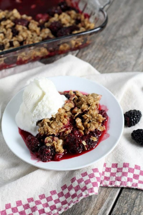 serving of blackberry crisp on a white plate with vanilla ice cream. Set on top of a white and red/white checked cloth napkin. Pan of crisp in the background + fresh blackberries