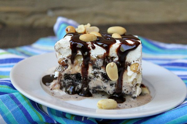 Slice of copycat Buster Bars on a white plate, topped with fudge and peanuts, displayed on a blue/teal/white striped napkin